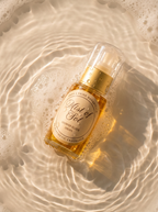 A bottle of Pride Botanicals Mist of Sol shower gel, filled with golden liquid and topped with a gold pump, resting on ripples of white foam in warm, sunlit water.