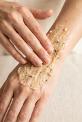 Close-up of a person's hands applying a textured, beige scrub with visible black specks to the back of another hand.